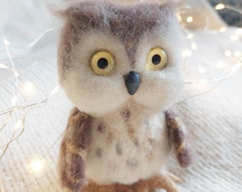 May include: A small, hand-felted owl with brown, white, and gray fur. The owl has large, yellow eyes and a black beak. The owl is sitting on a white, fuzzy surface.
