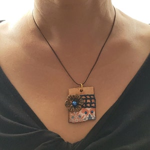 May include: A necklace with a rectangular pendant featuring a blue and orange floral design with a black crocheted trim. The pendant is made of ceramic and has a gold-coloured metal accent.