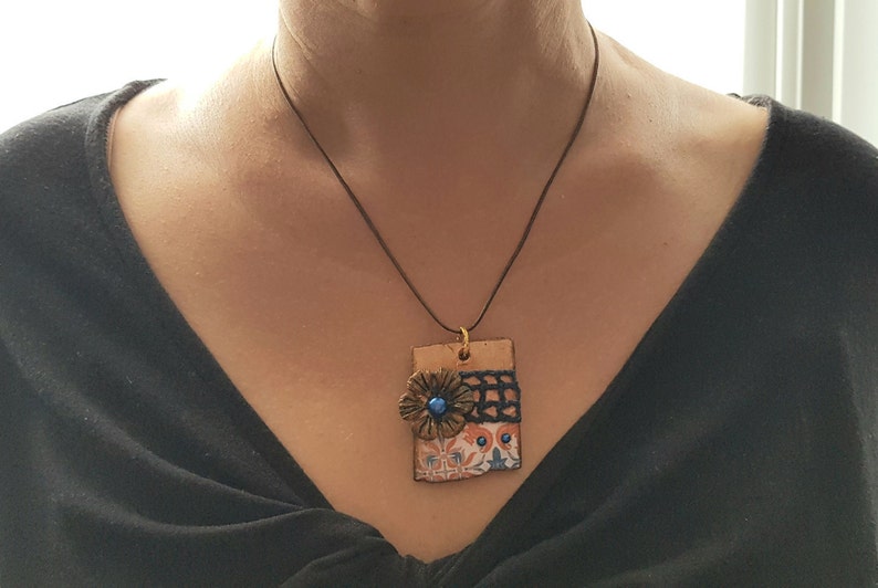 May include: A necklace with a rectangular pendant featuring a blue and orange floral design with a black crocheted trim. The pendant is made of ceramic and has a gold-coloured metal accent.