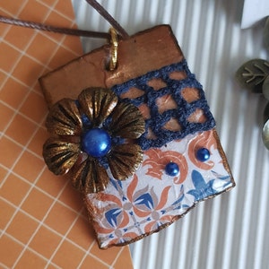May include: A copper-toned pendant with a blue and white patterned design, a gold-coloured flower with a blue gem in the centre, and a black lace overlay.