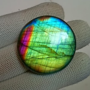 Puede incluir: Una piedra preciosa redonda de labradorita verde con destellos iridiscentes azules, morados y rojos. La piedra es lisa y pulida.