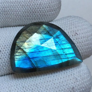 May include: A faceted, semi-circular labradorite gemstone. The stone displays a vibrant play of colour, with flashes of blue, teal, and gold. The cut reveals a geometric pattern, enhancing the stone's natural beauty. The gemstone is set against a white background.
