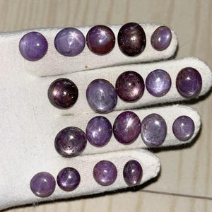 May include: A selection of polished, oval and round purple star sapphires. The gemstones vary in size and display a star-like effect. The sapphires are arranged on a white surface, showcasing their colour and lustre.