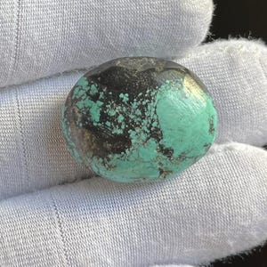 Può includere: Una pietra rotonda e lucidata con un mix di colori. La pietra presenta una sorprendente combinazione di nero, turchese e sfumature dorate. Le aree turchesi hanno un aspetto strutturato. La pietra è tenuta in un guanto bianco.