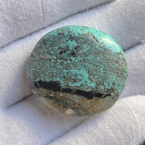 Puede incluir: Una piedra preciosa pulida y redonda con un llamativo patrón de color turquesa y negro. La piedra tiene una textura moteada con áreas de gris oscuro y marrón. La piedra preciosa tiene aproximadamente 5 cm de diámetro.