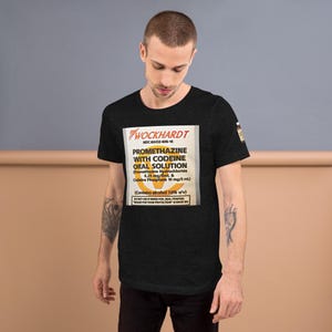 May include: Black t-shirt featuring a printed label design. The label reads "WOCKHARDT PROMETHAZINE WITH CODEINE ORAL SOLUTION" with details about the ingredients and alcohol content. The shirt is a short-sleeved crew neck.