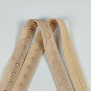 May include: Two beige bra hook and eye closures. One has multiple hook and eye sets, the other has a soft, fuzzy texture. The closures are made of a soft, flexible material.