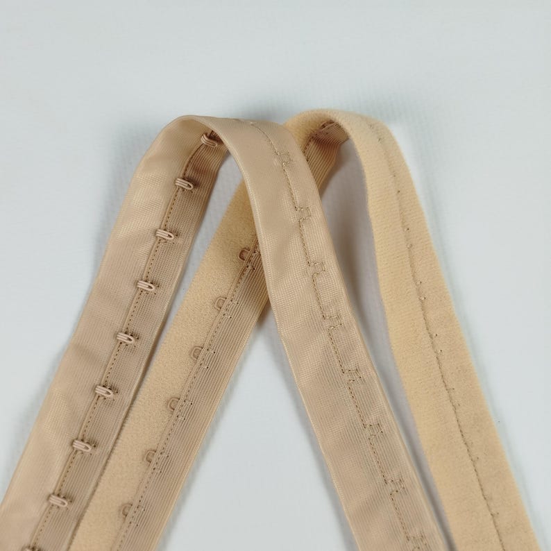 May include: Two beige bra hook and eye closures. One has multiple hook and eye sets, the other has a soft, fuzzy texture. The closures are made of a soft, flexible material.