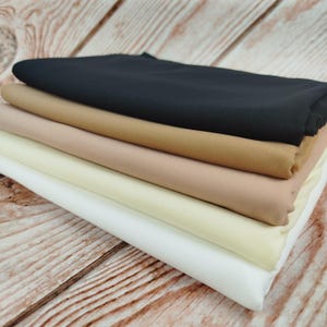 May include: A stack of fabric in various neutral colors: black, tan, beige, cream, and white. The fabric is neatly folded, showcasing the different shades. The fabrics are arranged on a wooden surface.
