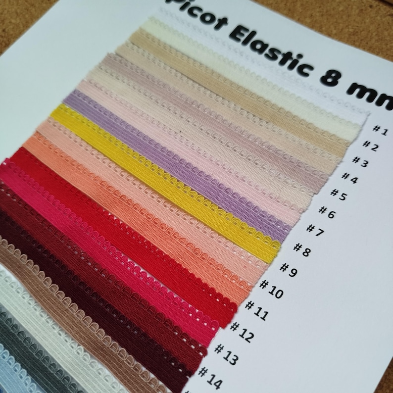 May include: A color chart of 14 different colors of 8mm picot elastic. The colors are arranged in rows, starting with white and ending with a dark blue. Each color is labeled with a number from 1 to 14.