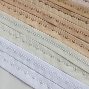 May include: Close-up of several rows of hook-and-eye closures in various neutral colours, including white, cream, beige, and tan. The closures are made of fabric and feature small metal hooks and eyes for fastening garments.