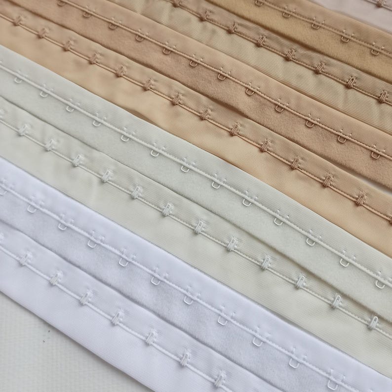 May include: Close-up of several rows of hook-and-eye closures in various neutral colours, including white, cream, beige, and tan. The closures are made of fabric and feature small metal hooks and eyes for fastening garments.