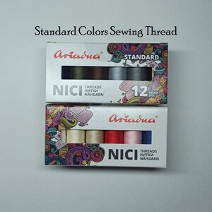 May include: Two boxes of Ariadna NICI sewing thread. The top box has six spools of thread in shades of gray, brown, and green. The bottom box has six spools of thread in shades of red, pink, beige, and blue. Both boxes have the text "Ariadna" and "NICI" on them.