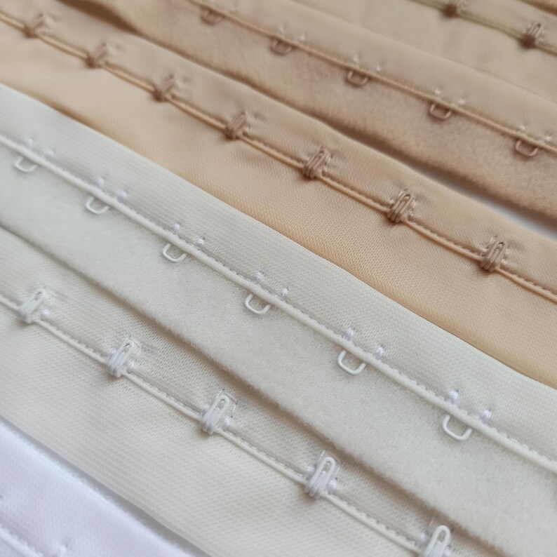 May include: Close-up of several rows of hook-and-eye closures in various neutral colours, including white, cream, and beige. The closures are made of fabric and metal, and are designed for use in garments.