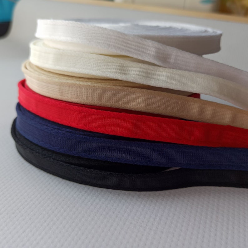 Puede incluir: Un mont&oacute;n de ribetes de tela de diferentes colores, incluyendo blanco, beige, rojo, azul marino y negro. El ribete est&aacute; doblado y apilado sobre una superficie blanca.