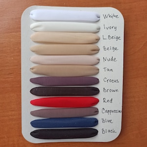 May include: A white display card featuring twelve fabric-covered elastic bands arranged horizontally. The colors are labeled as White, Ivory, L. Beige, Beige, Nude, Tan, Crocus, Brown, Red, Cappuccino, Blue, and Black in handwritten black ink.