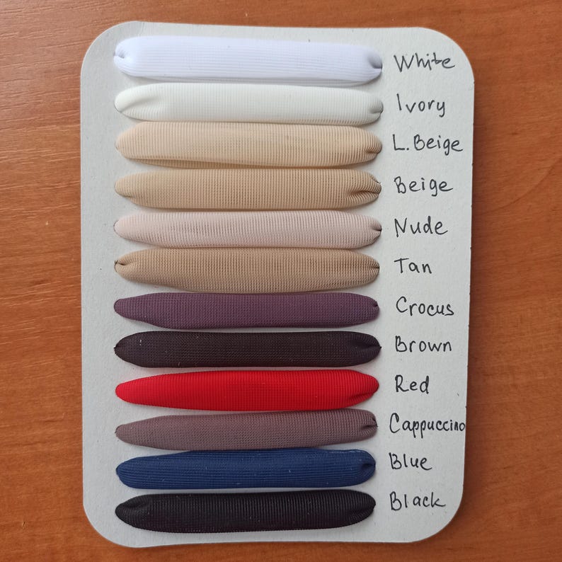 May include: A white display card featuring twelve fabric-covered elastic bands arranged horizontally. The colors are labeled as White, Ivory, L. Beige, Beige, Nude, Tan, Crocus, Brown, Red, Cappuccino, Blue, and Black in handwritten black ink.