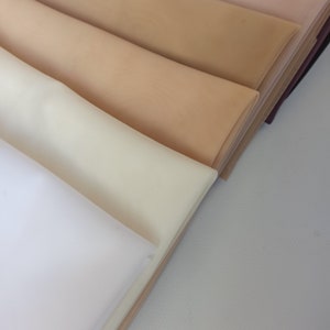 May include: A collection of folded fabric swatches arranged in a diagonal stack. The layers feature a range of neutral tones including white, cream, light beige, tan, and a darker brown shade, all displaying a fine, uniform ribbed texture.