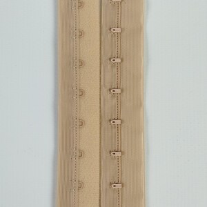 May include: A beige bra extender with hook-and-eye fastenings. The extender features multiple rows of hooks and eyes for adjustable sizing. The fabric is a solid beige colour, with visible stitching.