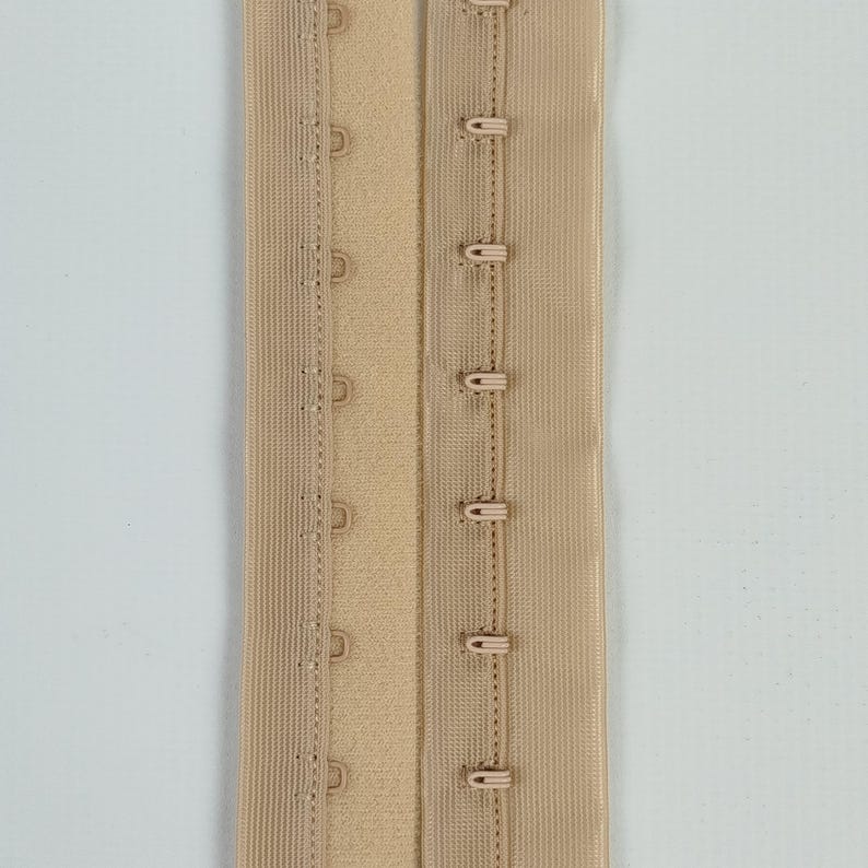 May include: A beige bra extender with hook-and-eye fastenings. The extender features multiple rows of hooks and eyes for adjustable sizing. The fabric is a solid beige colour, with visible stitching.