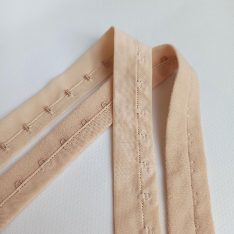 May include: Three beige bra hook and eye closures are arranged diagonally against a white background. The closures are made of fabric and feature multiple hook and eye sets for adjustable sizing. The fabric has a soft, matte finish.