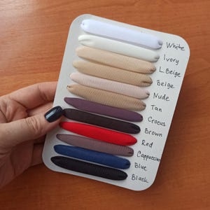 May include: A white display card featuring eleven horizontal fabric strips in various colors, labeled with handwritten text: White, Ivory, L. Beige, Beige, Nude, Tan, Crocus, Brown, Red, Cappuccino, Blue, and Black.
