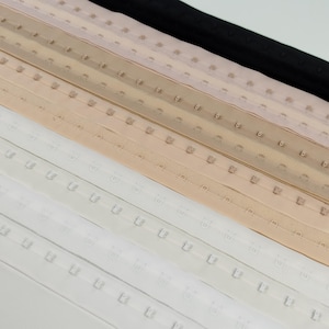 Peut inclure: Gros plan de plusieurs rangées de ruban à crochets et œillets de différentes couleurs. Les couleurs incluent le blanc, le beige, le beige clair, le rose clair et le noir. Le ruban à crochets et œillets est utilisé pour la couture et la confection de vêtements.