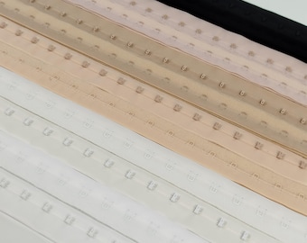 Könnte beinhalten: Nahaufnahme von mehreren Reihen Haken- und Ösenband in verschiedenen Farben. Die Farben umfassen Weiß, Beige, Hellbraun, Hellrosa und Schwarz. Das Haken- und Ösenband wird zum Nähen und für die Kleidungsherstellung verwendet.
