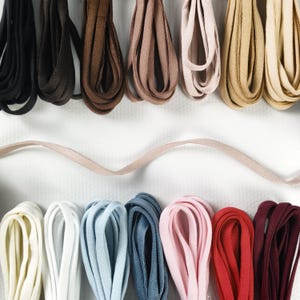 Pode incluir: Uma coleção de faixas elásticas planas dispostas em duas fileiras, apresentando uma variedade de cores incluindo preto, marrom, bege, branco, azul claro, azul escuro, rosa, vermelho e bordô, com uma faixa rosa claro esticada horizontalmente.