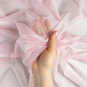 May include: A sheer, pale pink mesh fabric with a fine, semi-transparent texture. The lightweight material is gathered and draped, showing a soft, delicate appearance with consistent color throughout the entire piece of textile.