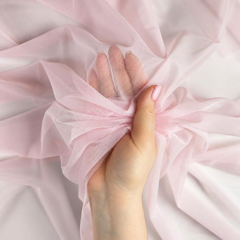 May include: A sheer, pale pink mesh fabric with a fine, semi-transparent texture. The lightweight material is gathered and draped, showing a soft, delicate appearance with consistent color throughout the entire piece of textile.