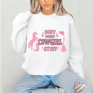 May include: White sweatshirt with a pink graphic design that says "Busy Doing Cowgirl Stuff". The graphic also includes a pink horse, cowboy hat, boot, and guitar.