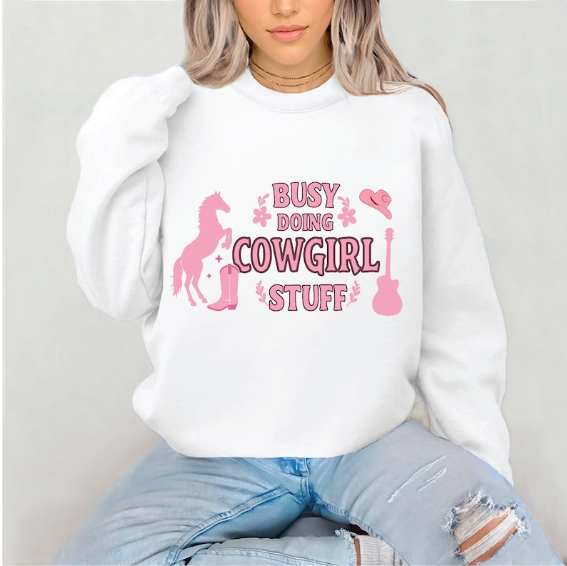 May include: White sweatshirt with a pink graphic design that says "Busy Doing Cowgirl Stuff". The graphic also includes a pink horse, cowboy hat, boot, and guitar.