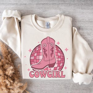 May include: A cream-colored sweatshirt with a pink cowboy boot graphic and a pink disco ball. The text "COWGIRL" is printed in pink below the graphic.