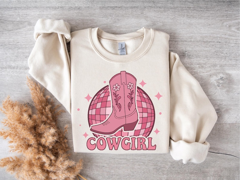 May include: A cream-colored sweatshirt with a pink cowboy boot graphic and a pink disco ball. The text "COWGIRL" is printed in pink below the graphic.
