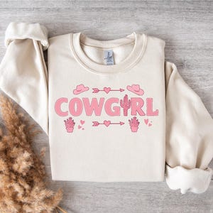 May include: A cream-colored sweatshirt with the text "COWGIRL" in pink letters. The design also includes pink cactus, hearts, and arrows.