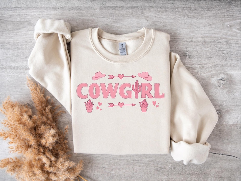 May include: A cream-colored sweatshirt with the text "COWGIRL" in pink letters. The design also includes pink cactus, hearts, and arrows.