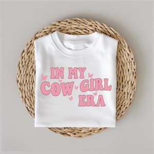 May include: White t-shirt with pink text that reads "In My Cowgirl Era" with pink butterfly graphics.