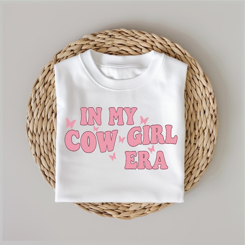 May include: White t-shirt with pink text that reads "In My Cowgirl Era" with pink butterfly graphics.