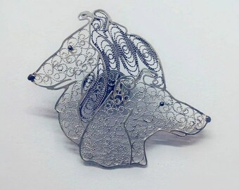 Puede incluir: Un broche de filigrana de plata con dos cabezas de perro entrelazadas. El broche está hecho de alambre de plata delicado y tiene un diseño detallado.