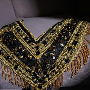 Puede incluir: Un collar negro y dorado con flecos. El collar tiene un escote en V y está decorado con lentejuelas y cuentas.