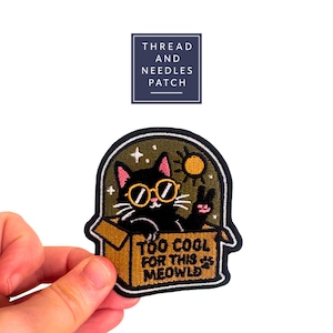 Könnte beinhalten: Gestickter Aufnäher mit einer schwarzen Katze mit Sonnenbrille, die ein Friedenszeichen gibt, in einer Pappschachtel. Der Aufnäher trägt den Text "TOO COOL FOR THIS MEOWLD" und hat einen dunkelgrünen Rand. Der Aufnäher ist von "THREAD AND NEEDLES PATCH".