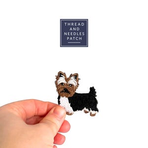 Può includere: Toppa ricamata di un cane Yorkshire Terrier con pelo marrone, nero e bianco. La toppa è tenuta da una mano su sfondo bianco. Il testo "THREAD AND NEEDLES PATCH" è in un quadrato blu navy.