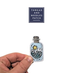 May include: Embroidered patch depicting a bottle with a nautical theme. The bottle design includes a sun, waves, and clouds in blue, white, and yellow. The patch is held against a white backdrop. The text "THREAD AND NEEDLES PATCH" is in a navy blue box.