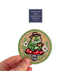 Può includere: Una toppa rotonda ricamata con una rana verde con cappello e tazza, seduta su un fungo rosso a pois bianchi. La toppa ha un bordo beige con dettagli floreali e la scritta "THREAD AND NEEDLES PATCH".