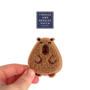 Peut inclure: Un écusson brodé marron représentant un capybara de dessin animé avec un corps texturé et de petits bras. L'écusson a une bordure marron clair et des détails marron foncé. Le texte "THREAD AND NEEDLES PATCH" est dans un rectangle bleu marine.