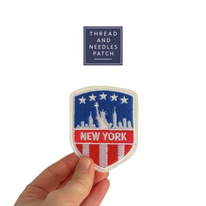 May include: A shield-shaped embroidered patch featuring a New York City design. The patch has a blue top with five white stars, a white skyline, and red and white stripes at the bottom. The word "NEW YORK" is in red. The patch is held by a hand.