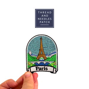 Puede incluir: Parche bordado con la Torre Eiffel en París. El parche tiene un fondo de cielo azul, follaje verde y una pancarta blanca con la palabra "París". El parche tiene forma de arco. También es visible el texto "THREAD AND NEEDLES PATCH".