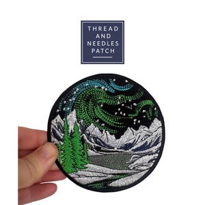 May include: Embroidered patch featuring a mountain landscape under a starry night sky with green and blue aurora borealis. The patch is round and has the text "THREAD AND NEEDLES PATCH" in a navy blue square.