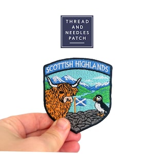 Può includere: Toppa ricamata con un design delle Highlands scozzesi. La toppa include una mucca delle Highlands, una pulcinella di mare e il testo "Scottish Highlands" in blu. La toppa è tenuta in mano su sfondo bianco.